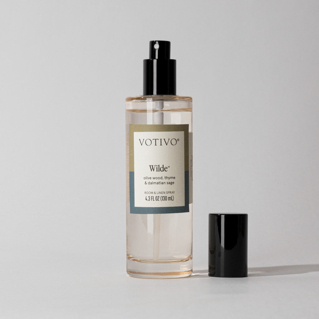 A clear Votivo Wilde Room & Linen Spray bottle—Olive Wood, Thyme & Damask Rose scent—with a black nozzle and cap beside it rests on a light gray surface. Refresh fabrics with this spray for lasting freshness.