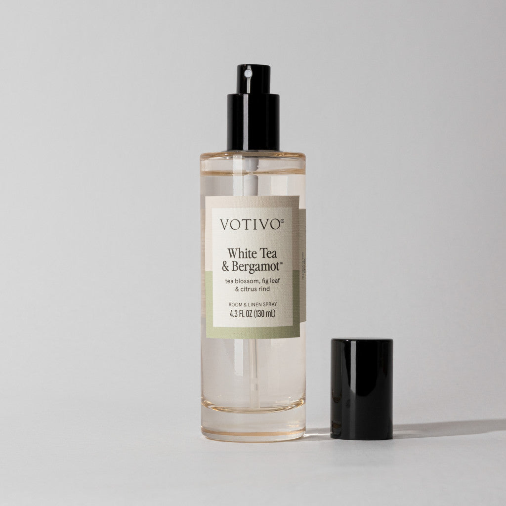 A clear Votivo White Tea & Bergamot Room & Linen Spray bottle with a black nozzle and fabric-safe black cap sits on a plain light gray background.