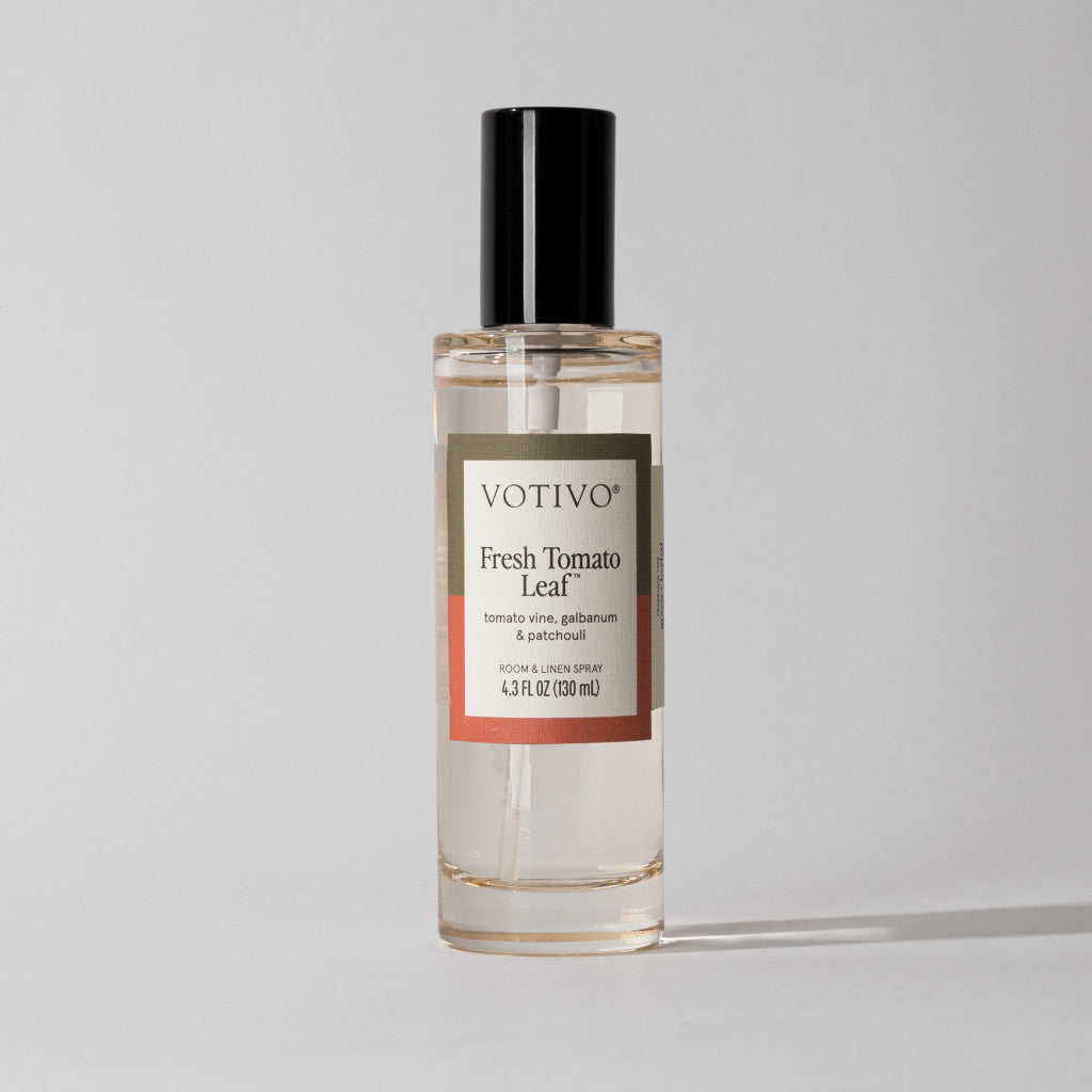 A clear Votivo Fresh Tomato Leaf Room & Linen Spray bottle with a black cap sits on a light background. The label features tomato vine, galbanum, and patchouli fragrance notes and indicates the spray is fabric safe.