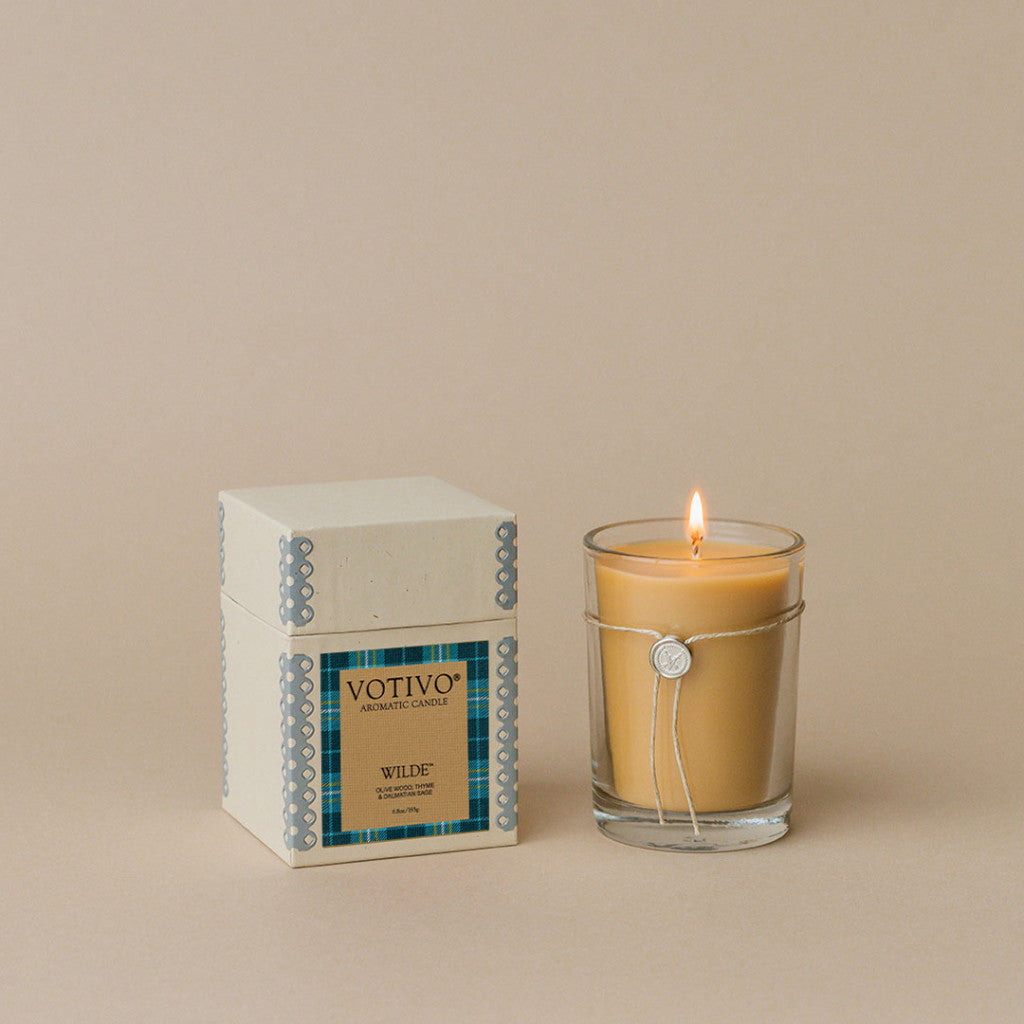 The Votivo Wilde 6.8 oz. Aromatic Candle, in a clear glass jar with a silver seal, sits beside its matching box on a beige background—enjoy its unique fragrance and soothing glow.