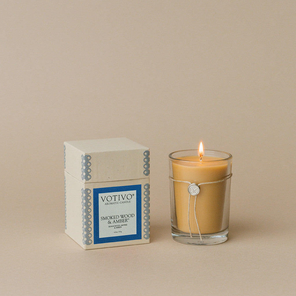 A lit beige Votivo Smoked Wood & Amber 6.8 oz. Aromatic Candle in a clear glass holder sits beside its box, made with soy blend wax, against a plain beige background.