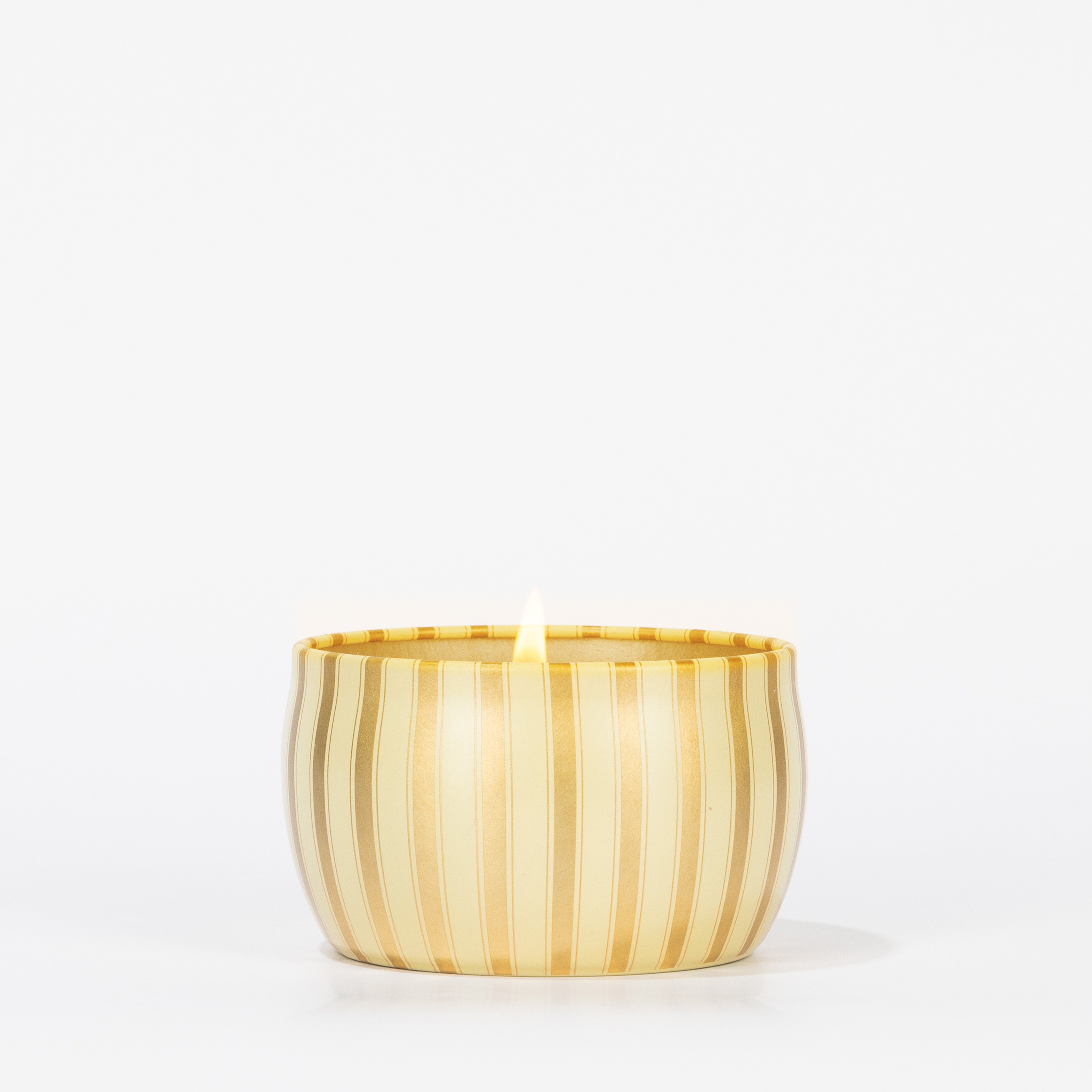 A single lit Votivo Gilded Pear Holiday Tin Candle, in a round gold tin with vertical stripes, is set against a plain white background.