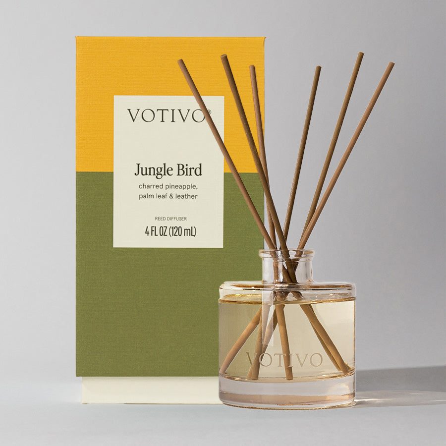 The Votivo Jungle Bird Reed Diffuser, with fiber reeds in a clear glass bottle, stands before its yellow and green box and fills your space with continuous notes of charred pineapple, palm leaf, and leather.