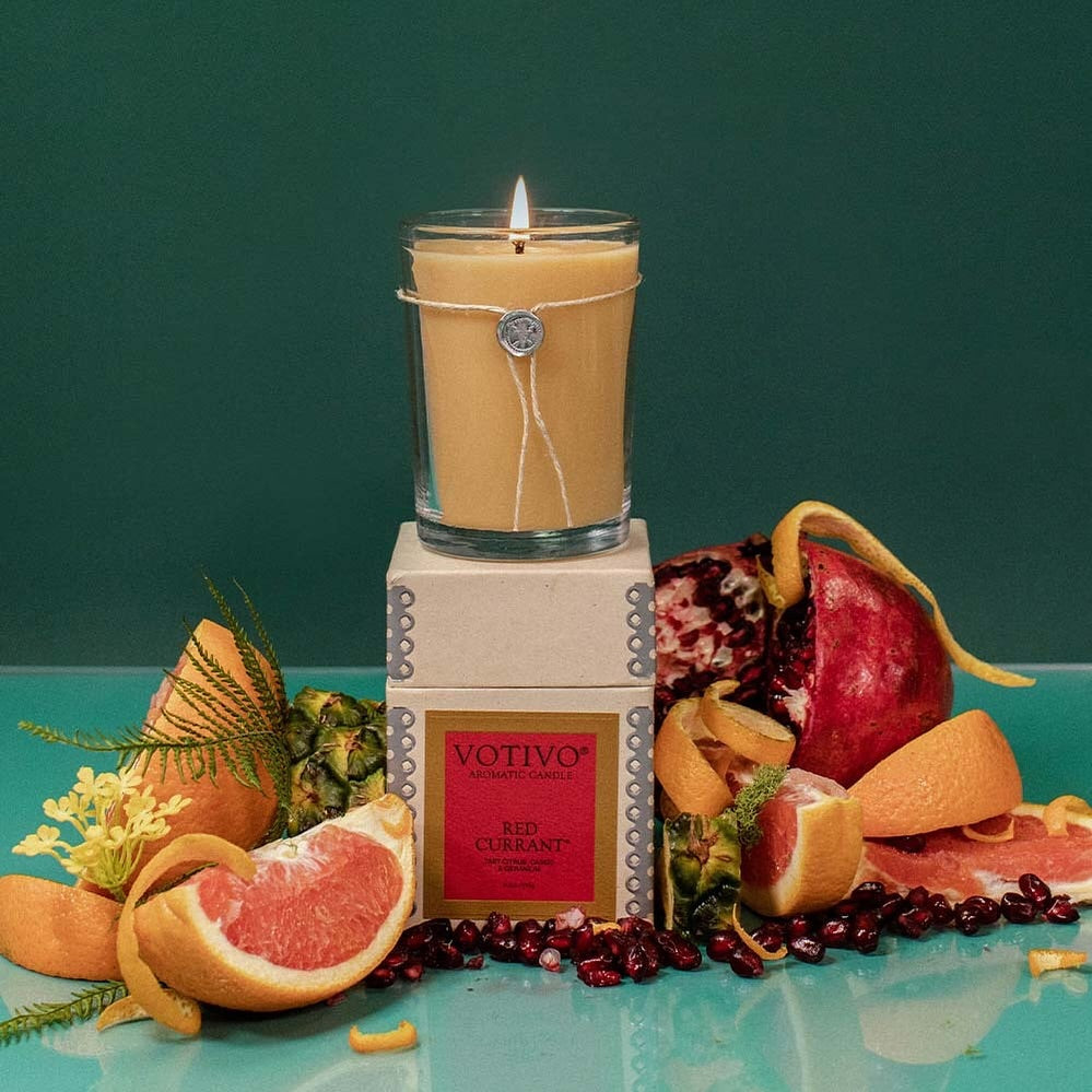 The Votivo Red Currant 6.8 oz. Aromatic Candle sits atop its box, surrounded by grapefruit slices, pomegranate, orange peels, and green leaves on a teal background. Its vibrant red currant scent fills the air.