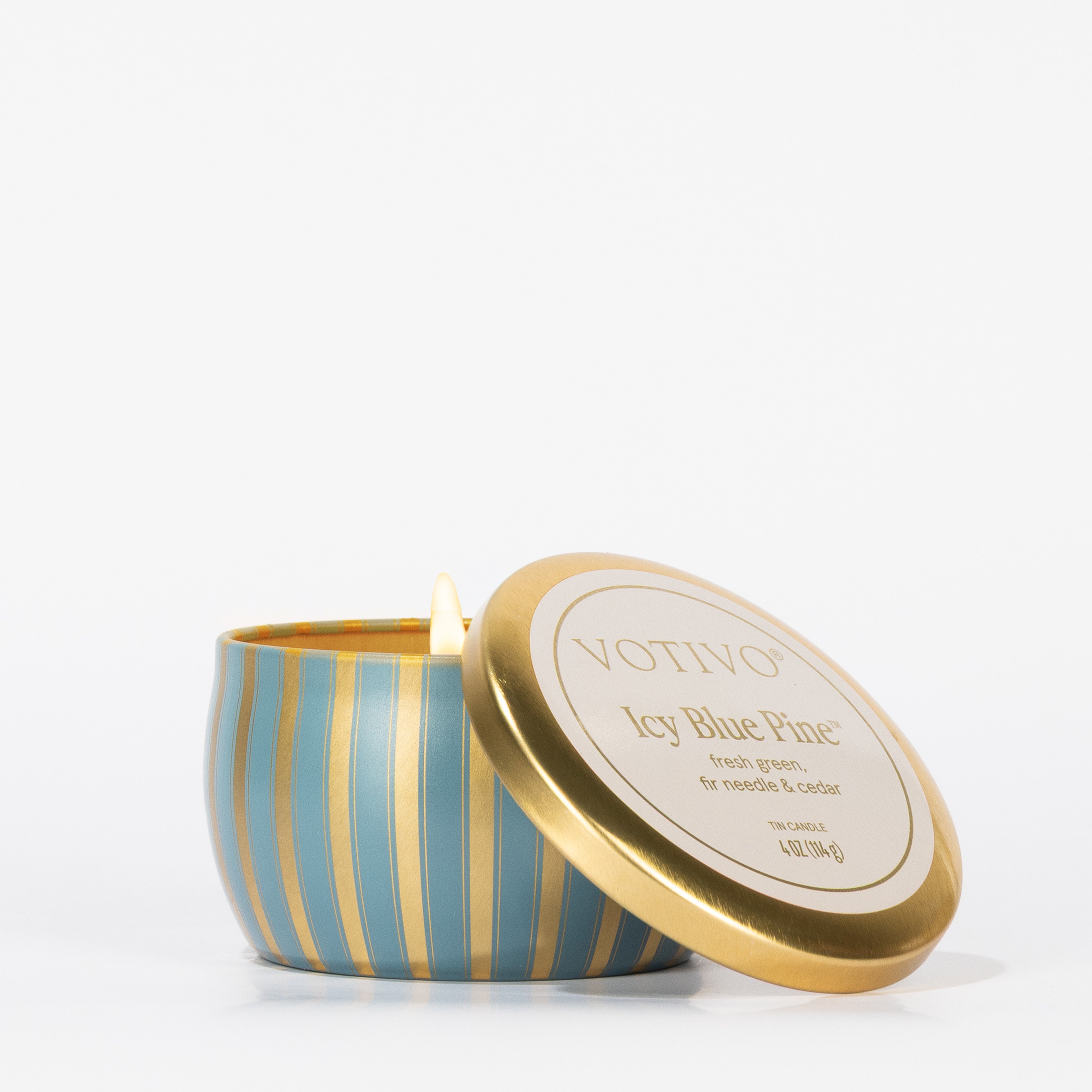 A lit Votivo Icy Blue Pine Holiday Tin Candle crafted with soy blend wax sits in a blue and gold striped tin, the lid off to the side, on a white background.
