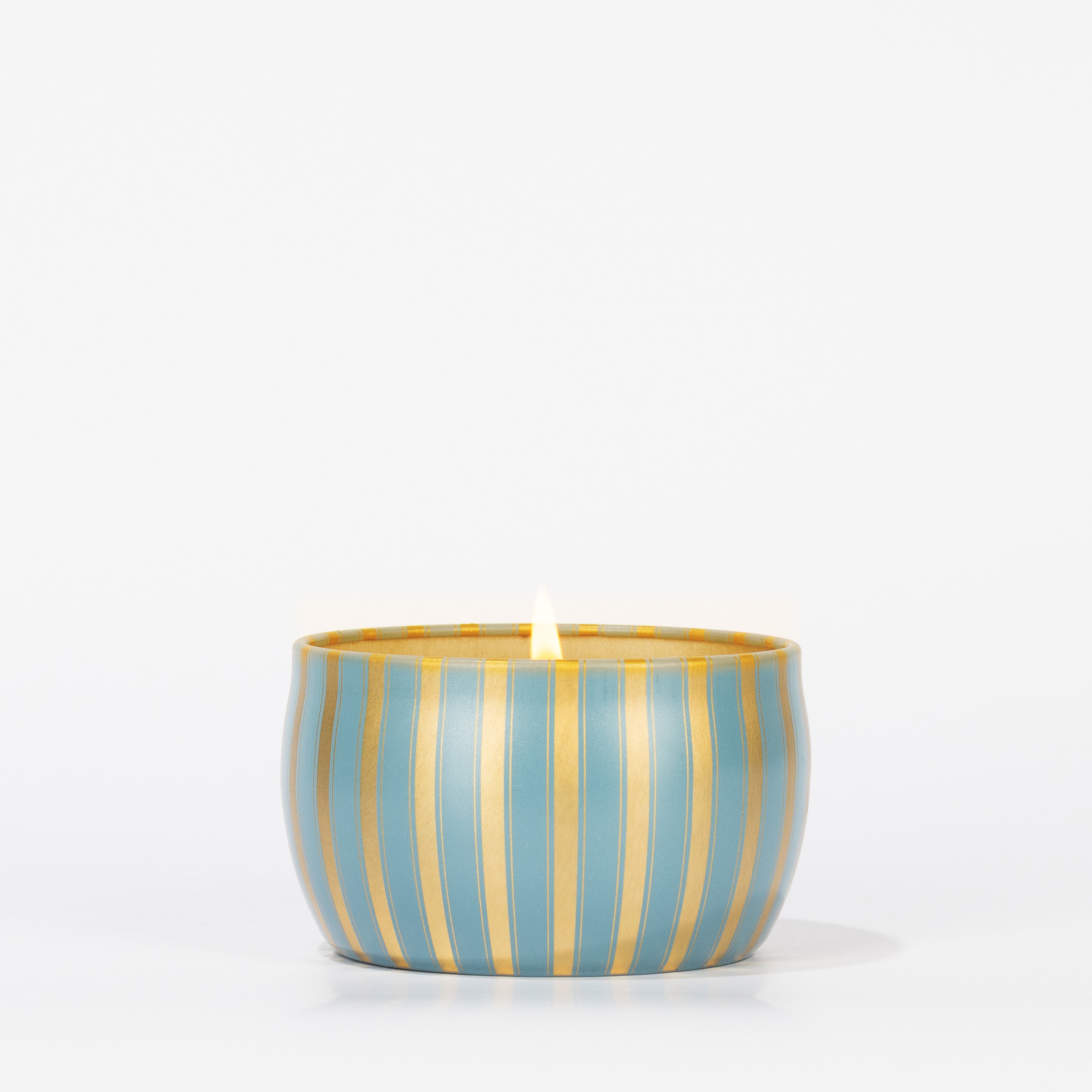 The Votivo Icy Blue Pine Holiday Tin Candle, in a round blue and gold striped container filled with soy blend wax, is displayed centered on a plain white background.