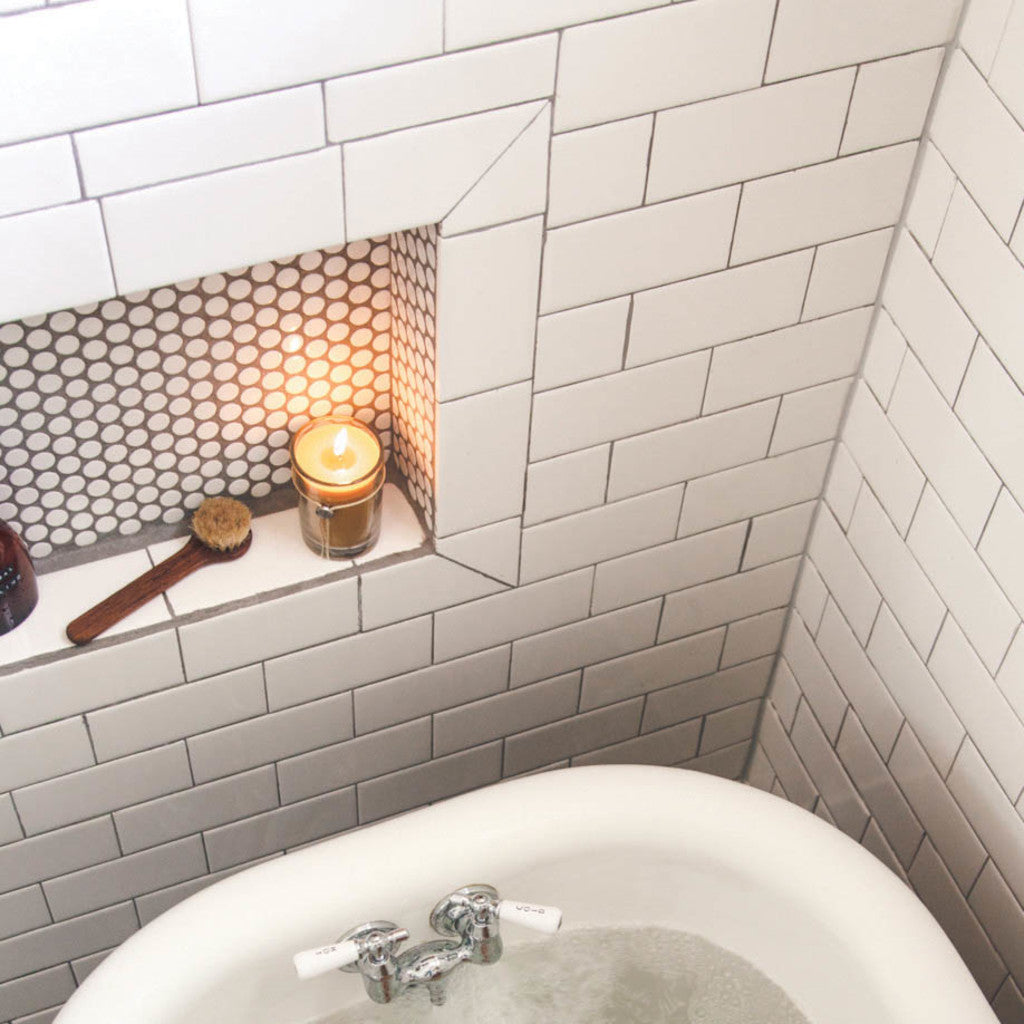 A white tiled bathroom features a built-in shelf with hexagonal tile accents, holding a Votivo Saint Germain Lavender 6.8 oz. Aromatic Candle, a brush, and a bottle above a partially filled white bathtub.