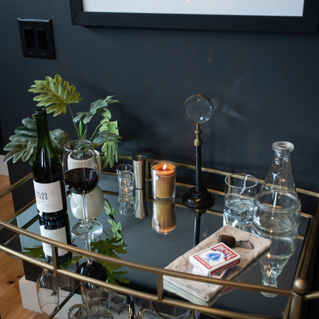 A gold and glass bar cart holds a wine bottle, filled glass, Votivo Smoked Wood & Amber 6.8 oz. Aromatic Candle, magnifying glass, leafy plant, water carafe, glasses, napkins, and playing cards against a dark wall.