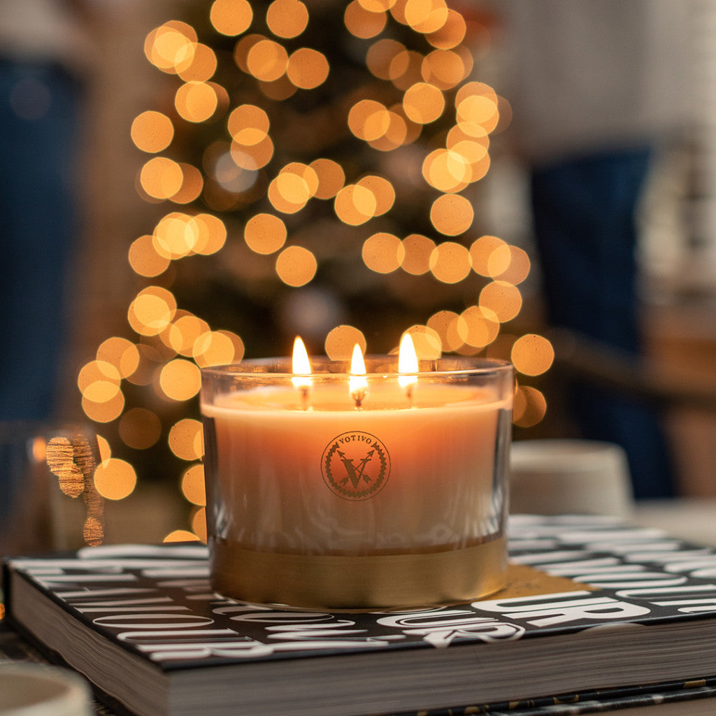 The Votivo Red Currant Holiday 3 Wick Candle glows in its glass holder atop a stack of books, surrounded by twinkling lights and a festive tree, filling the space with its signature red currant fragrance for a cozy holiday vibe.