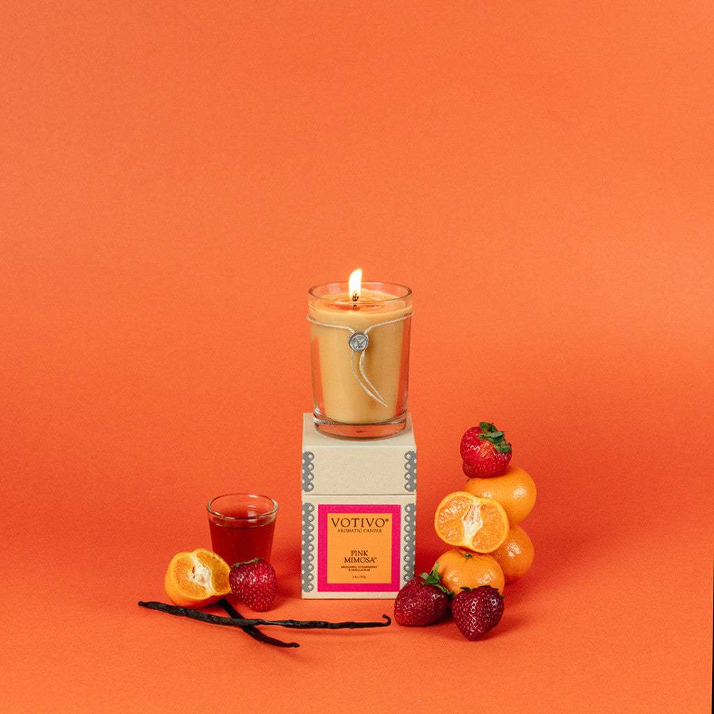 A Votivo Pink Mimosa 6.8 oz. Aromatic Candle sits on its box, surrounded by strawberries, tangerines, vanilla beans, and a glass of red liquid. Set against an orange background. Made with soy blend wax for a clean burn.