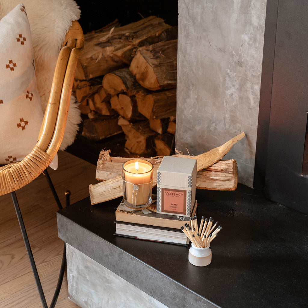 The Votivo Teak 6.8 oz. Aromatic Candle rests on stacked books beside its box, with matches nearby; firewood and part of a cushioned chair appear next to a cozy fireplace in the background.