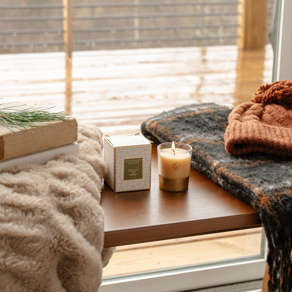 A Votivo Sequoia Fir Holiday Votive, cozy blanket, knit hat, and book sit on a wooden table by a rainy window, creating a warm atmosphere as the subtle scent of sequoia fir fills the air.