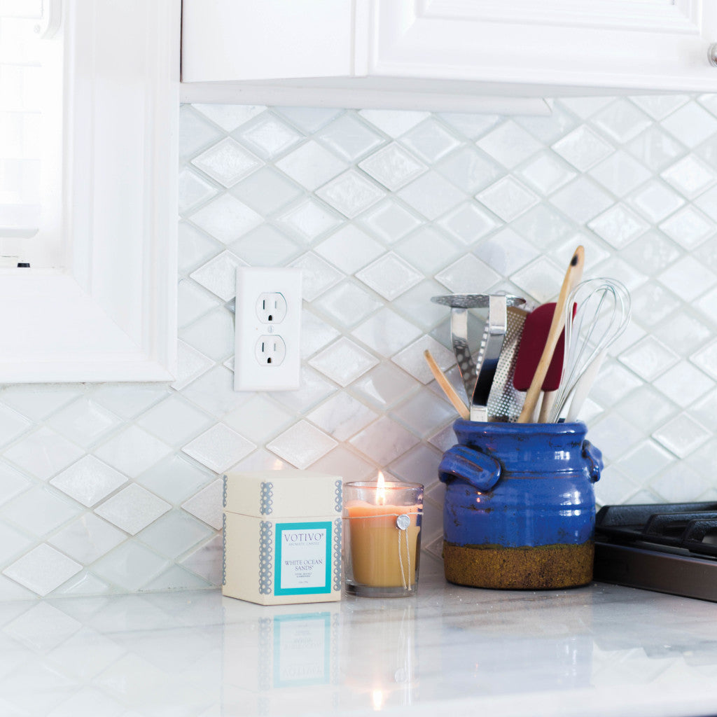 A kitchen counter displays a blue ceramic utensil holder with cooking tools, a Votivo White Ocean Sands 6.8 oz. aromatic candle, and a white box, all set against a white tile backsplash.