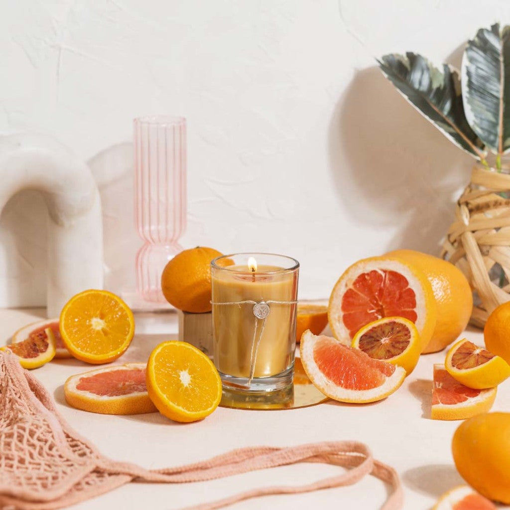 The Votivo Island Grapefruit 6.8 oz. Aromatic Candle, crafted with a soy blend wax, sits in a glass jar on a table amid sliced island grapefruit, a mesh bag, pink glass, and decorative basket with leaves against a white arch backdrop.