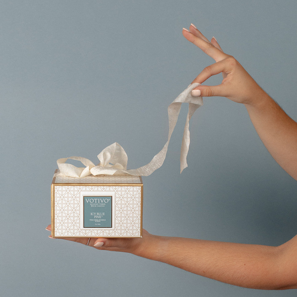 A person holds a Votivo Icy Blue Pine Holiday 3 Wick Candle box in one hand and pulls white tissue paper from the top, set against a plain gray background.