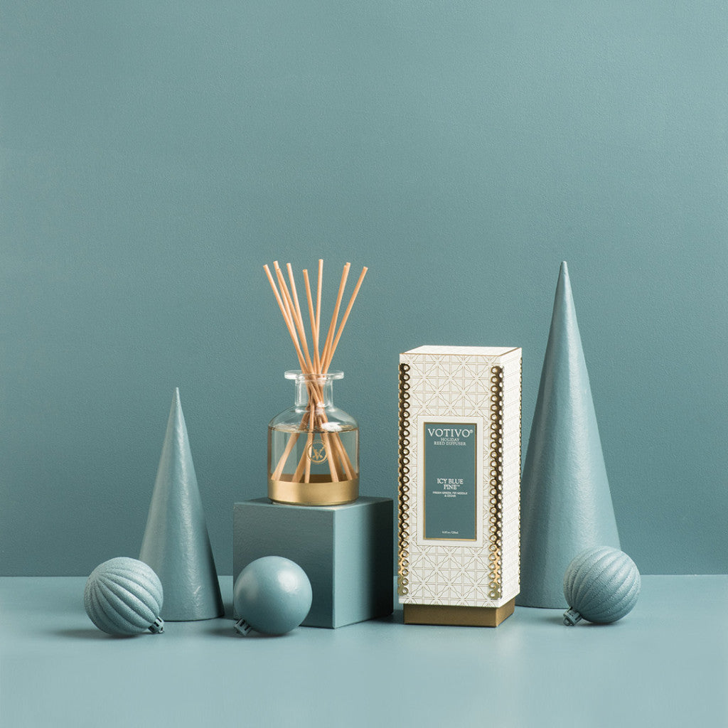 The Votivo Icy Blue Pine Holiday Reed Diffuser, with gold holder, reeds, and decorative box, is displayed among blue geometric shapes and ornaments, filling the scene with a fresh pine scent against a blue background.