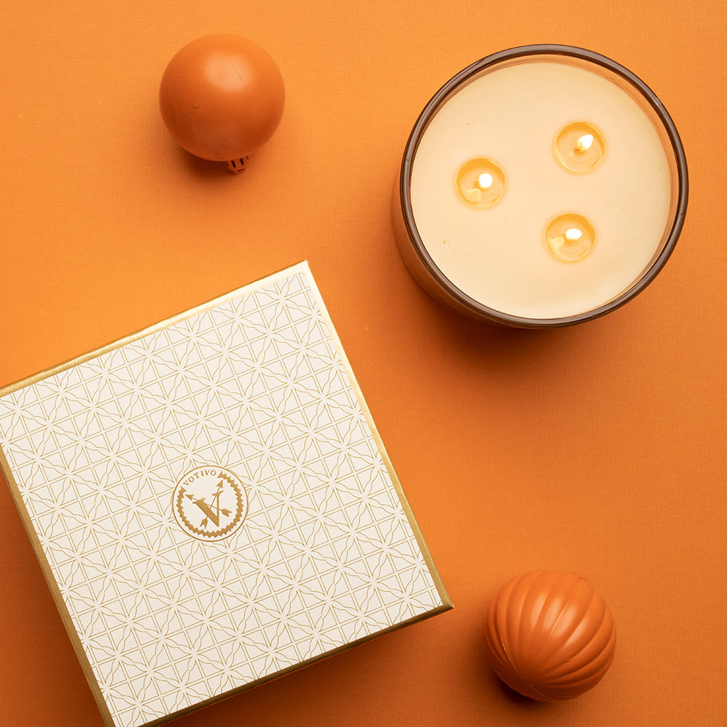 A lit Votivo Spiced Chai Holiday 3 Wick Candle with soy blend wax, a patterned white and gold Votivo box, and two orange decorative balls are arranged on an orange background.