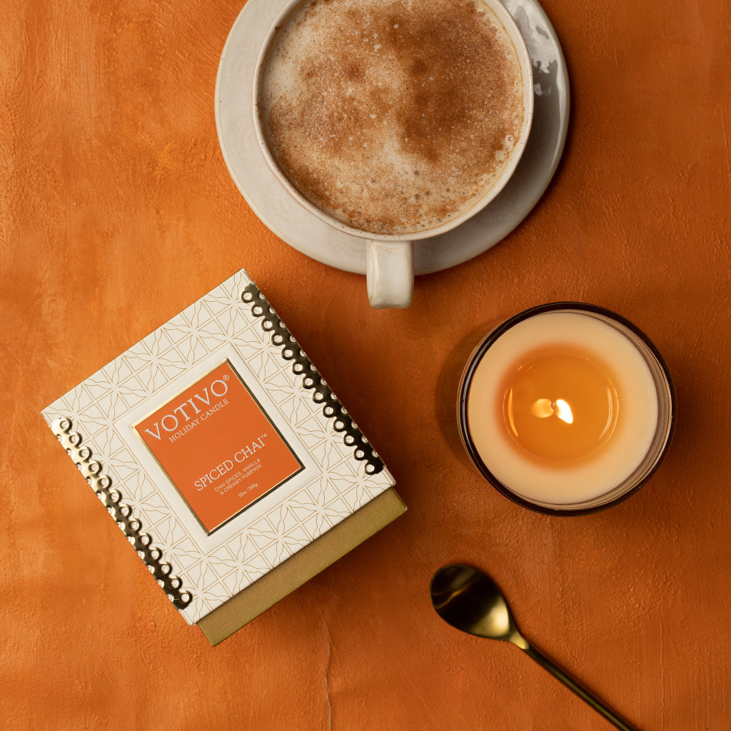 A cup of frothy chai latte, a lit Votivo Spiced Chai 10 oz. Holiday Candle in a glass holder, a gold spoon, and its festive box are artfully arranged on an orange surface.