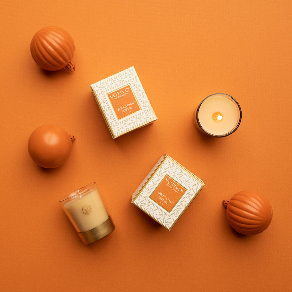 Overhead view of two Votivo Spiced Chai Holiday Votive boxes, two lit soy blend wax candles in glass holders, and three round orange ornaments arranged on an orange background.