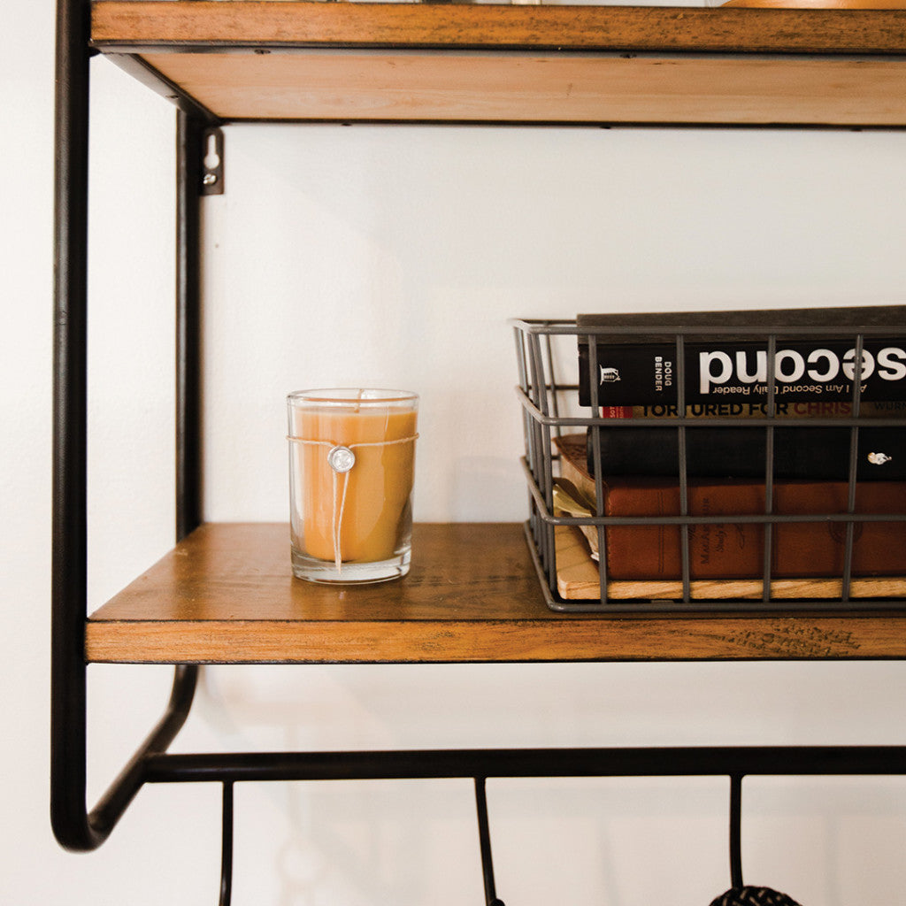A wooden shelf holds several books in a wire basket and a Votivo Champaca 6.8 oz. Aromatic Candle, all set against a white wall.