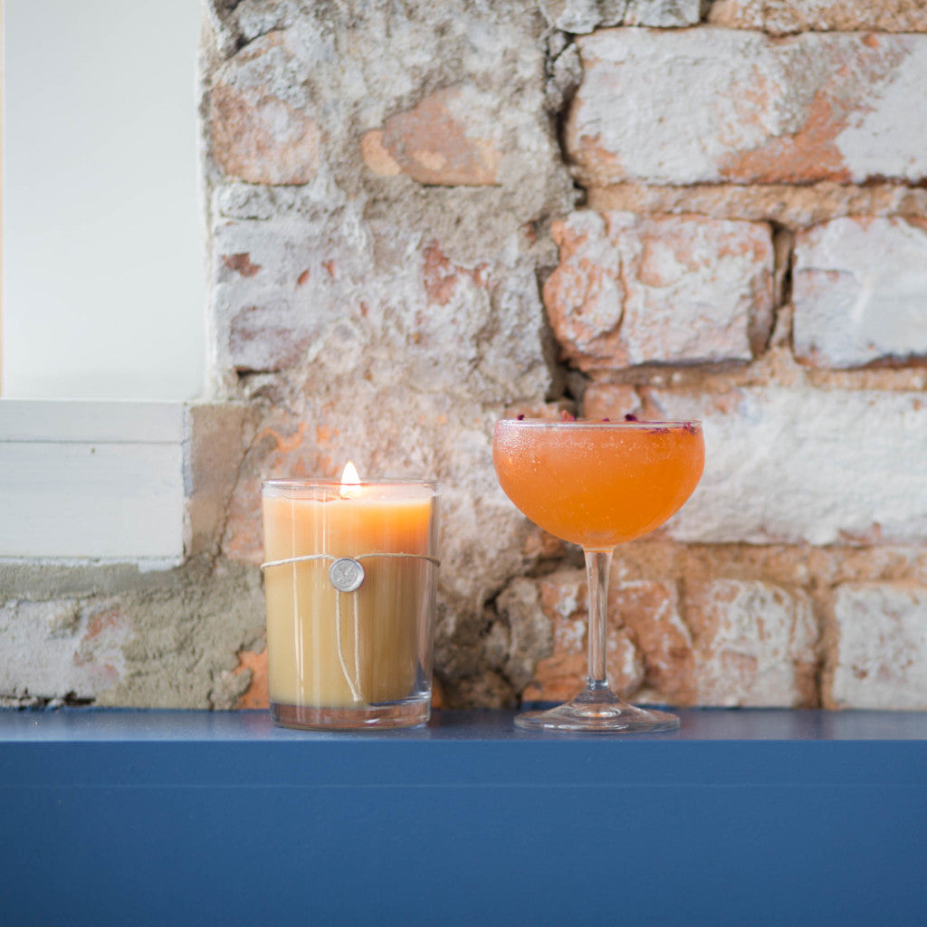 The Votivo Pink Mimosa 6.8 oz. Aromatic Candle in a glass jar and an orange cocktail rest on a blue ledge before a rustic brick wall, setting the scene for relaxation.