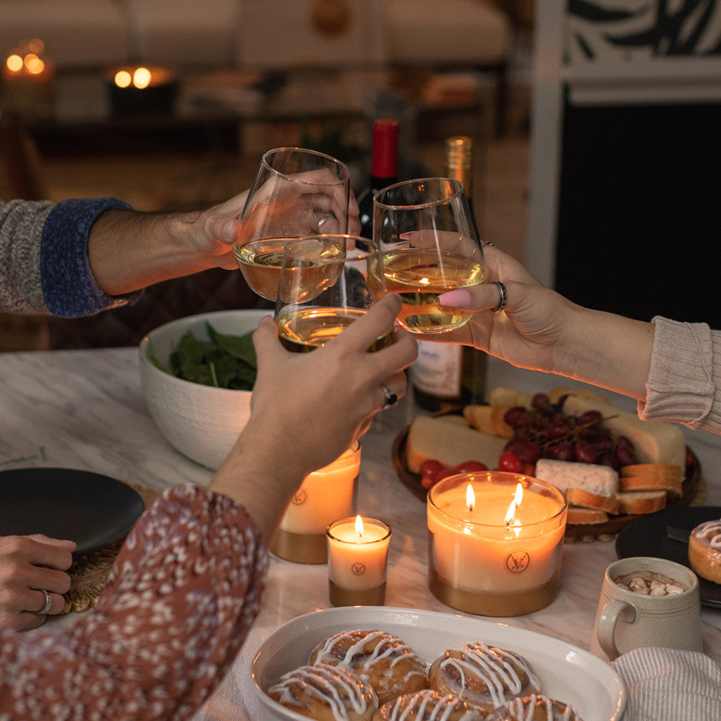 Three friends toast with white wine over a table of pastries, cheese, grapes, and salad, as the Votivo Red Currant Holiday 3 Wick Candle fills the cozy room with festive fragrance for a joyful celebration.