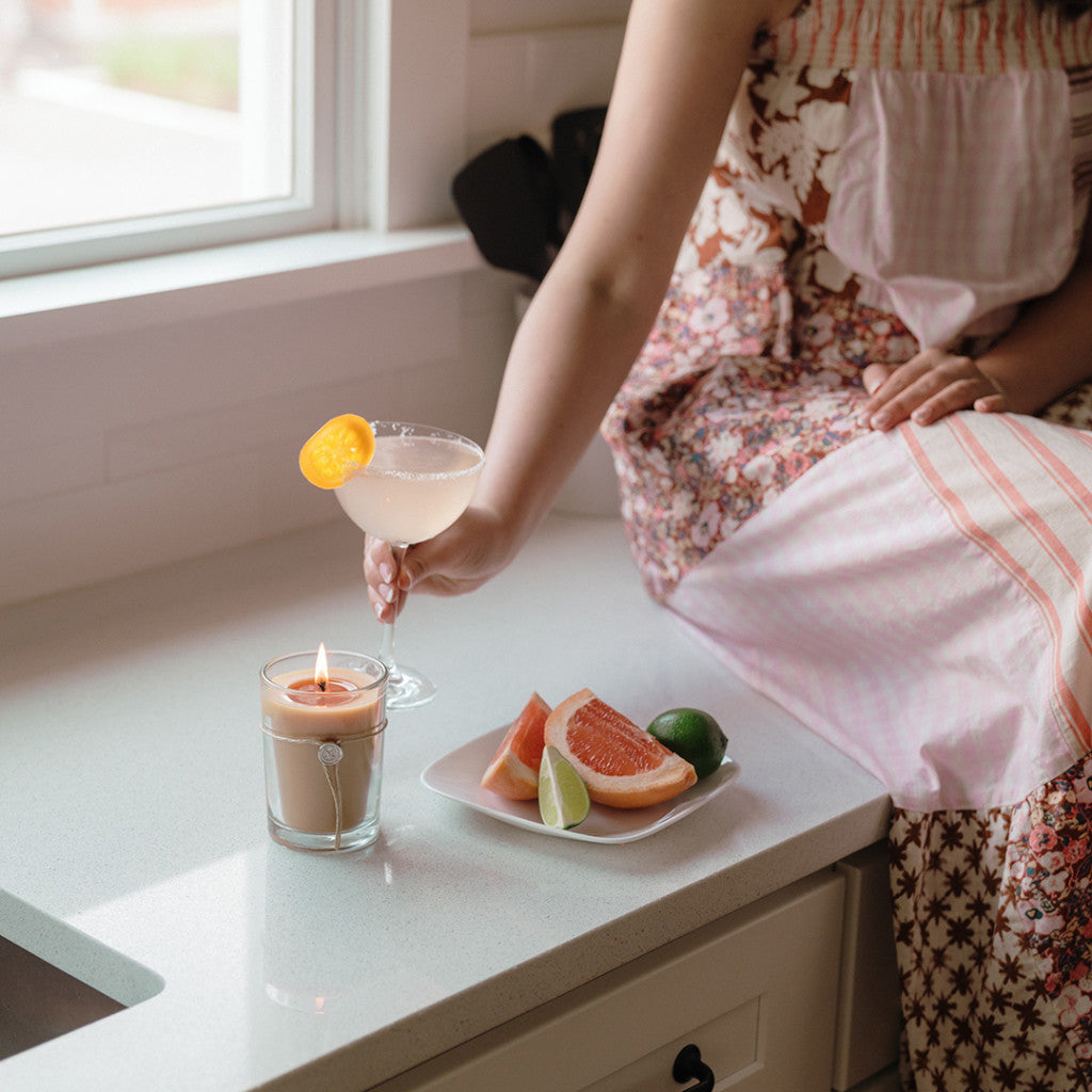 A person in a floral dress sits on a kitchen counter with a citrus cocktail, while the Votivo Island Grapefruit 6.8 oz. Aromatic Candle flickers nearby beside a plate of grapefruit wedges and lime.