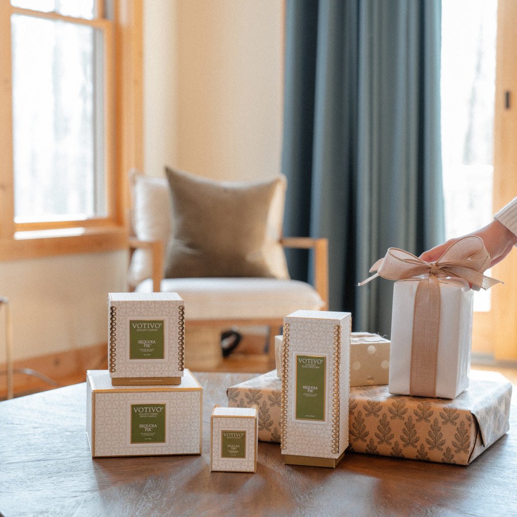 A hand sets a gift with a beige ribbon on a table among other presents, Votivo candles, and the Sequoia Fir Holiday Reed Diffuser by Votivo with Rattan Reeds, in a cozy living room featuring a chair and window.