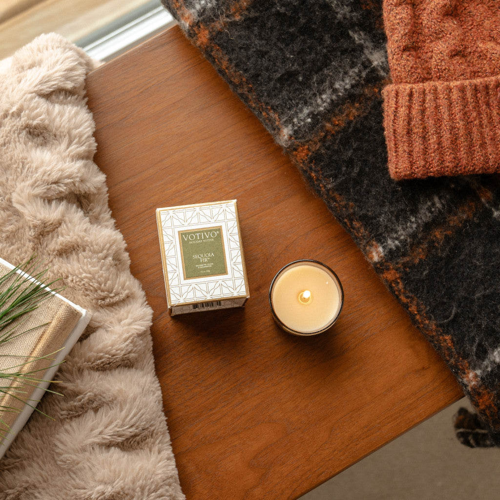 A lit Votivo Sequoia Fir Holiday Votive candle sits on a wooden surface next to a furry beige blanket, a black-and-brown plaid scarf, and a rust-colored knit hat.