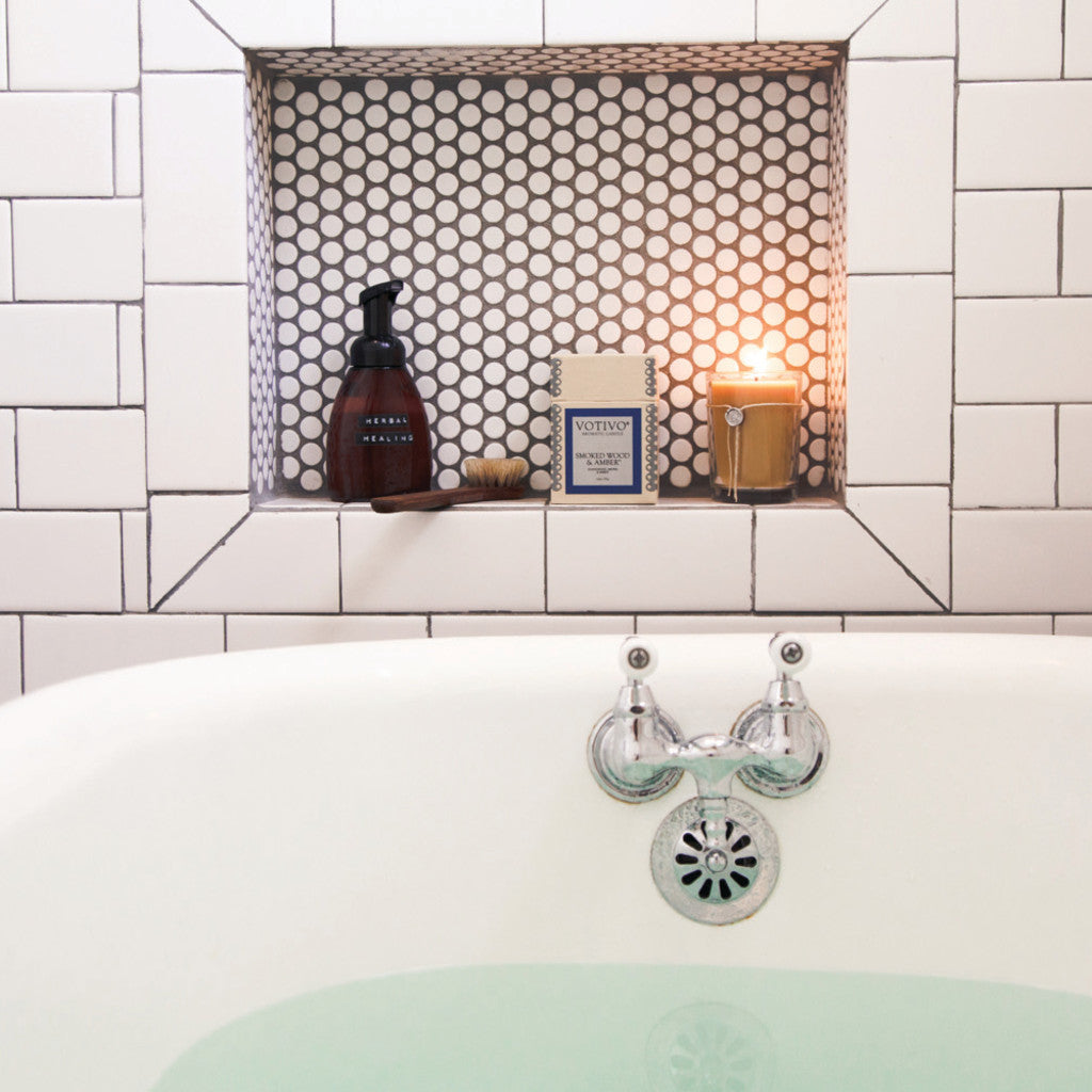 A white tiled bathtub filled with water sits beneath a recessed shelf holding a soap dispenser, box of soap, small brush, and a Votivo Smoked Wood & Amber 6.8 oz. Aromatic Candle, all set against hexagonal tiles.