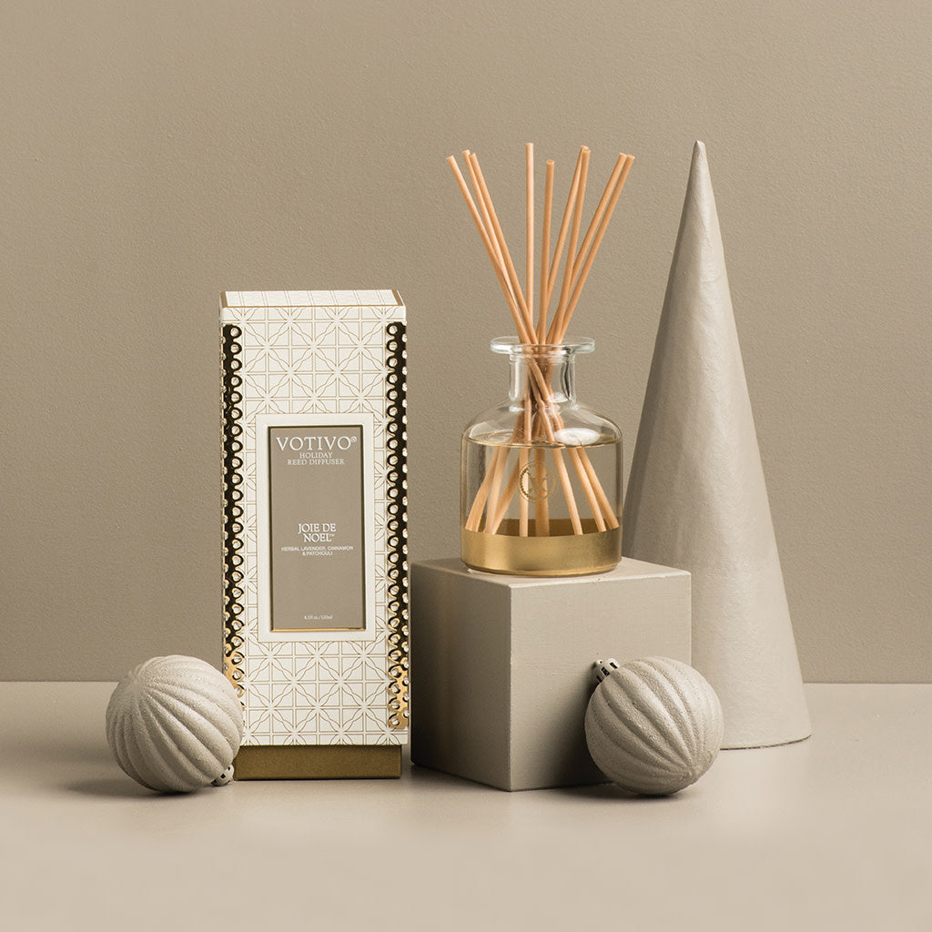 The Votivo Joie de Noel Holiday Reed Diffuser, in gold and white packaging, sits beside a glass diffuser with rattan reeds and textured gray ornaments on a neutral background, evoking festive holiday fragrance.