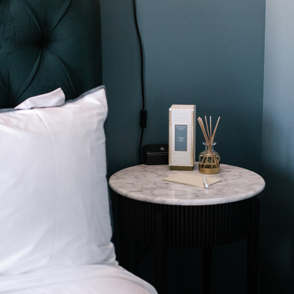 A marble-topped nightstand beside a bed with white pillows displays the Votivo Icy Blue Pine Holiday Reed Diffuser, a decorative box, a note, and an electronic device, set against a soothing blue wall.
