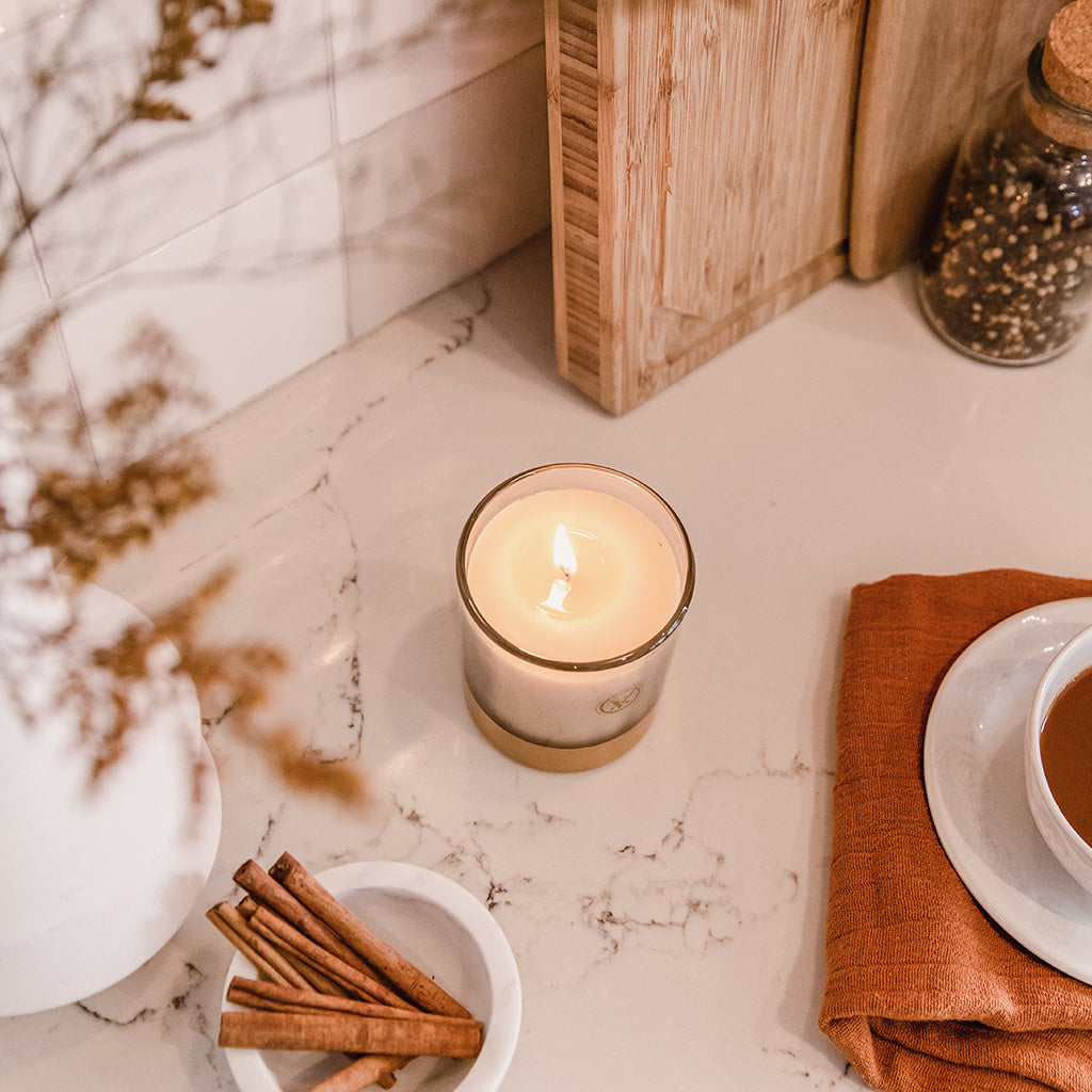 The Votivo Spiced Chai 10 oz. Holiday Candle, made with soy blend wax, glows in a glass jar on a marble countertop, surrounded by cinnamon sticks, an orange napkin, cutting boards, and dried flowers in the corner.