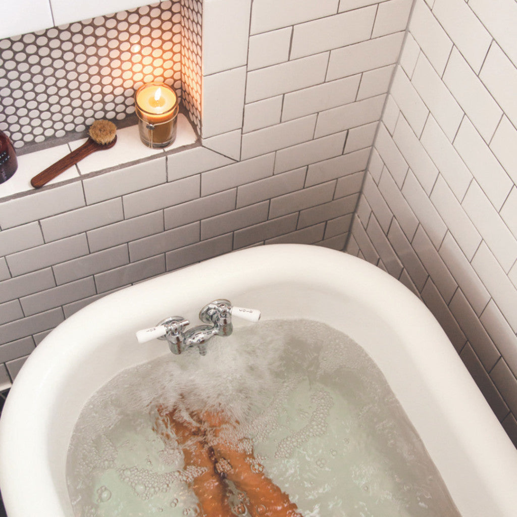 A person relaxes in a white bathtub with water, legs partially visible. A Votivo Clean Crisp White 6.8 oz. Aromatic Candle, brush, and small items sit on a tiled shelf above the tub. The walls are lined with white subway tiles.
