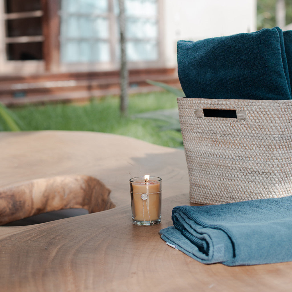 The Votivo White Ocean Sands 6.8 oz. Aromatic Candle in a glass holder rests on a wooden table beside a blue towel and basket, with greenery and large windows in the background, creating a serene coastal atmosphere.