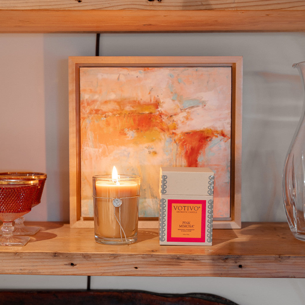 A Votivo Pink Mimosa 6.8 oz. Aromatic Candle sits in its box on a wooden shelf next to an orange glass container, framed by an abstract painting and a clear glass vase.