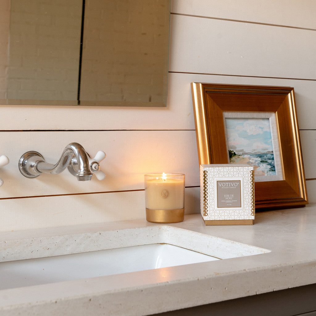 A Votivo Joie de Noel 10 oz. Holiday Candle and a decorative framed picture sit on a bathroom counter by the sink and mirror, bringing festive charm to the shiplap wall space.