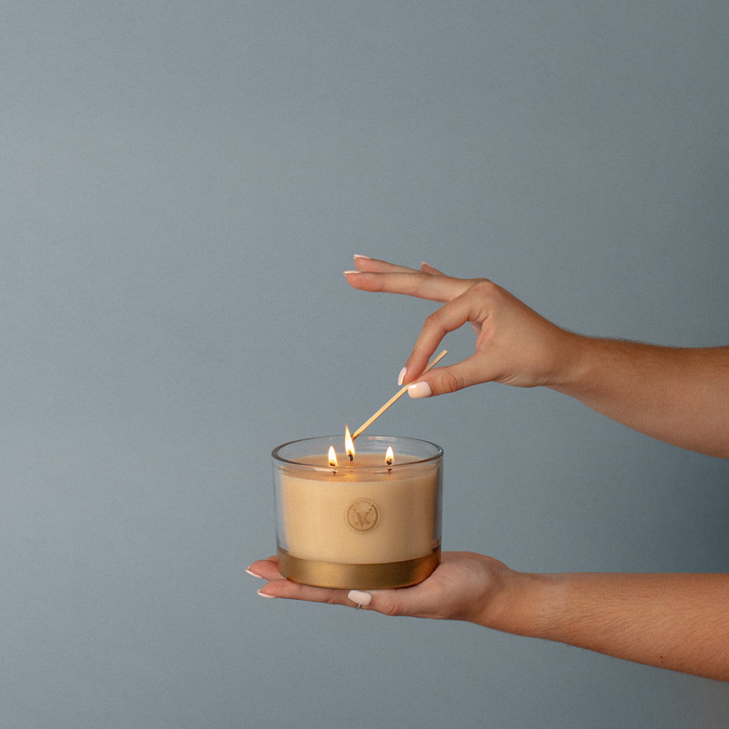 A person holds a Votivo Icy Blue Pine Holiday 3 Wick Candle, made from soy blend wax, in one hand and lights a wick with a match against a plain gray background.