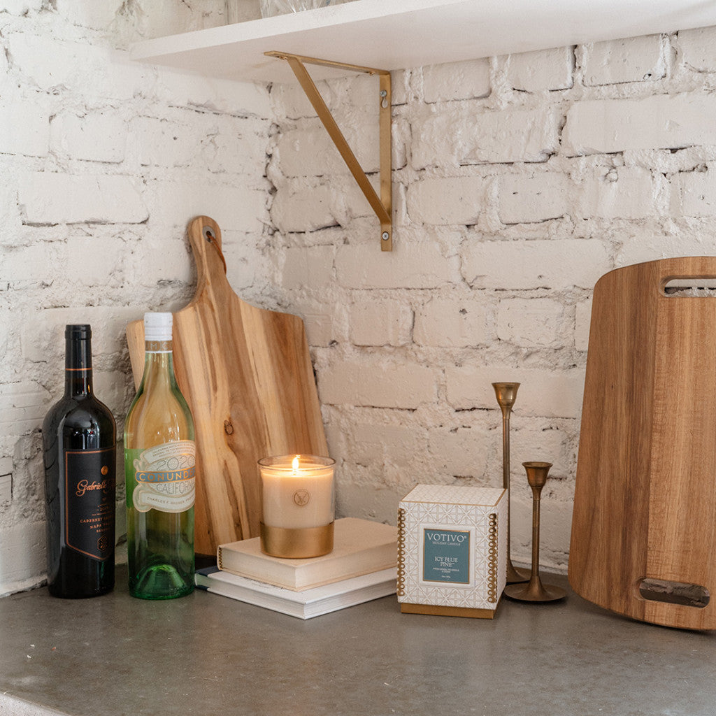 A kitchen countertop displays a wine bottle, green glass bottle, Votivo’s Icy Blue Pine 10 oz. Holiday Candle on books, a decorative candle box, wooden cutting boards, and brass candlesticks against a white brick wall.