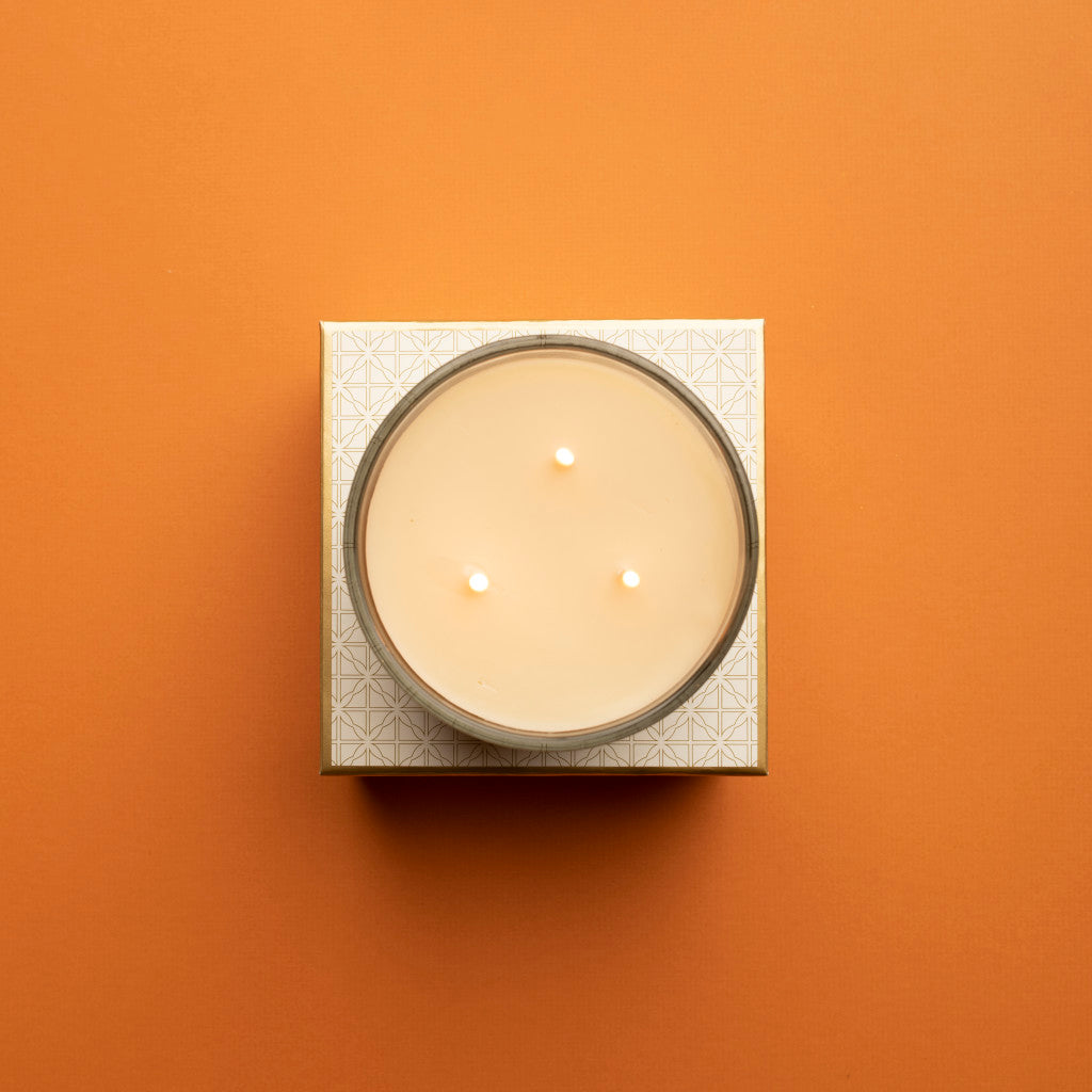 A top-down view of the Votivo Spiced Chai Holiday 3 Wick Candle, made with soy blend wax in a round container on a patterned square base, set against an orange background.