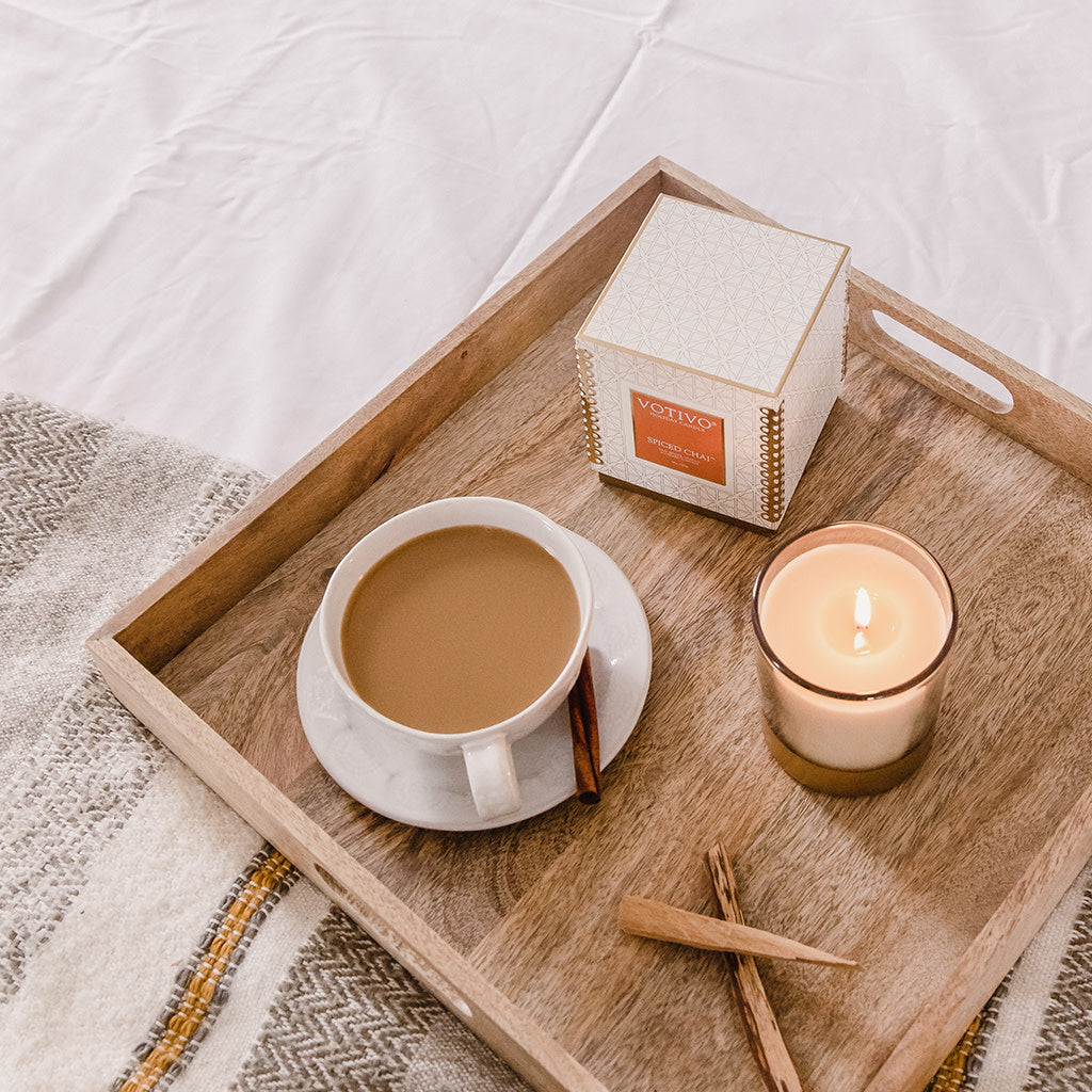 A wooden tray on a bed holds a cup of coffee with cinnamon, the Votivo Spiced Chai 10 oz. Holiday Candle made from soy blend wax, a Votivo box, and two cinnamon sticks, with a cozy blanket partially visible underneath.