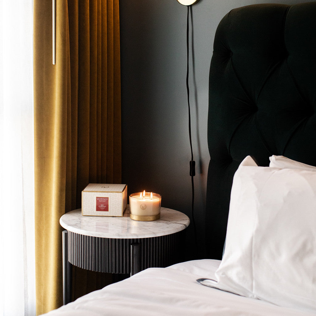 A Votivo Red Currant Holiday 3 Wick Candle sits on a round marble nightstand beside a neatly made bed with white sheets, gold curtains, and a dark upholstered headboard.