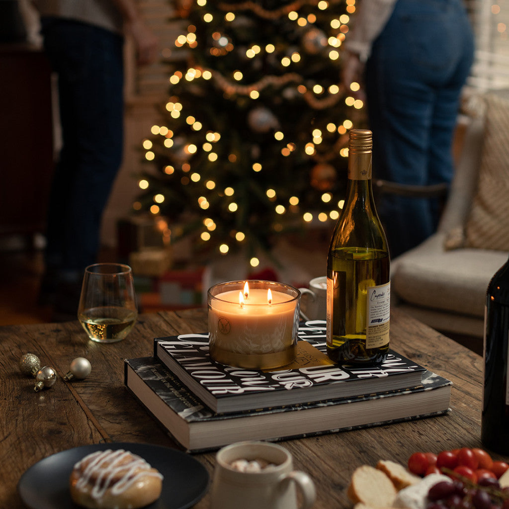 A cozy living room glows with a Votivo Red Currant Holiday 3 Wick Candle, its festive scent filling the air. Wine, books, and snacks rest on a wooden table beside a decorated Christmas tree as two people stand in the soft light.