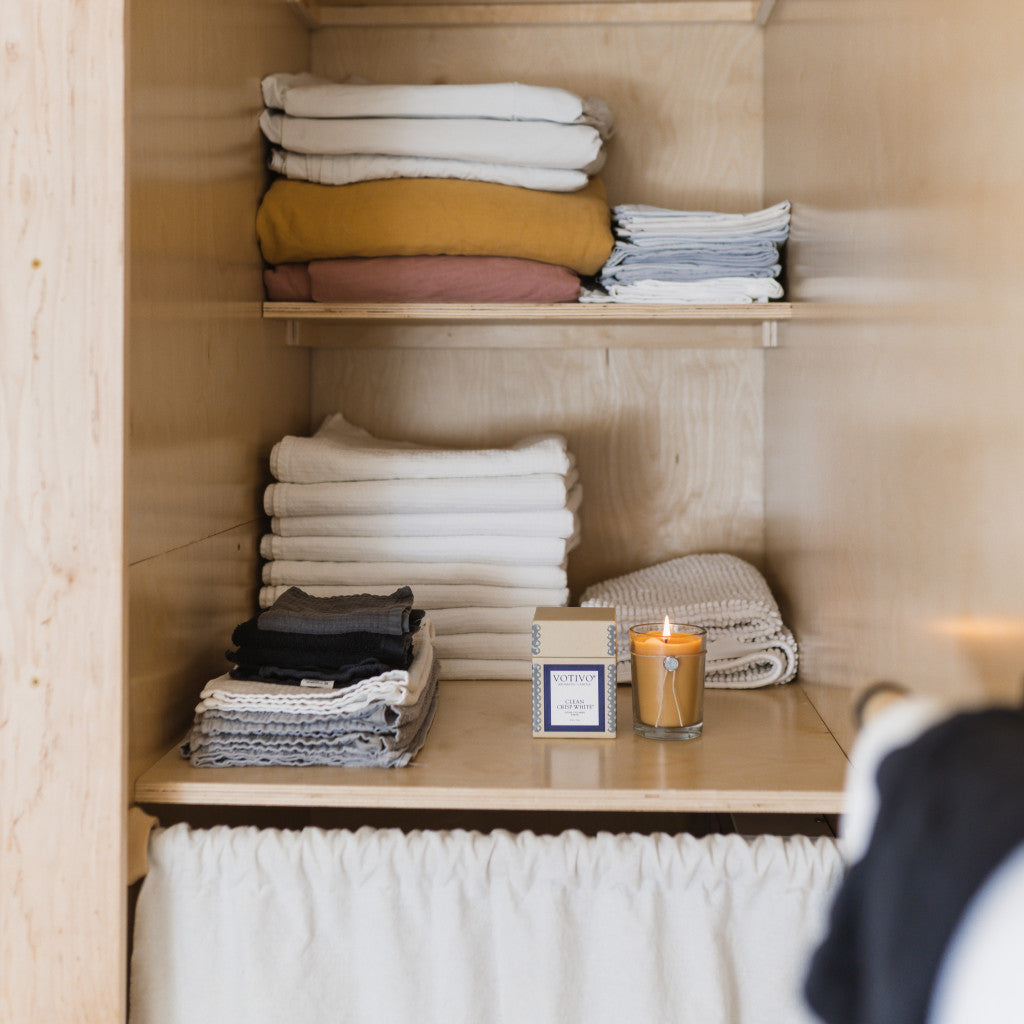 A wooden shelf with neatly folded white towels and linens is styled with a Votivo Clean Crisp White 6.8 oz. Aromatic Candle and a box, creating a cozy, minimalistic, and organized look.