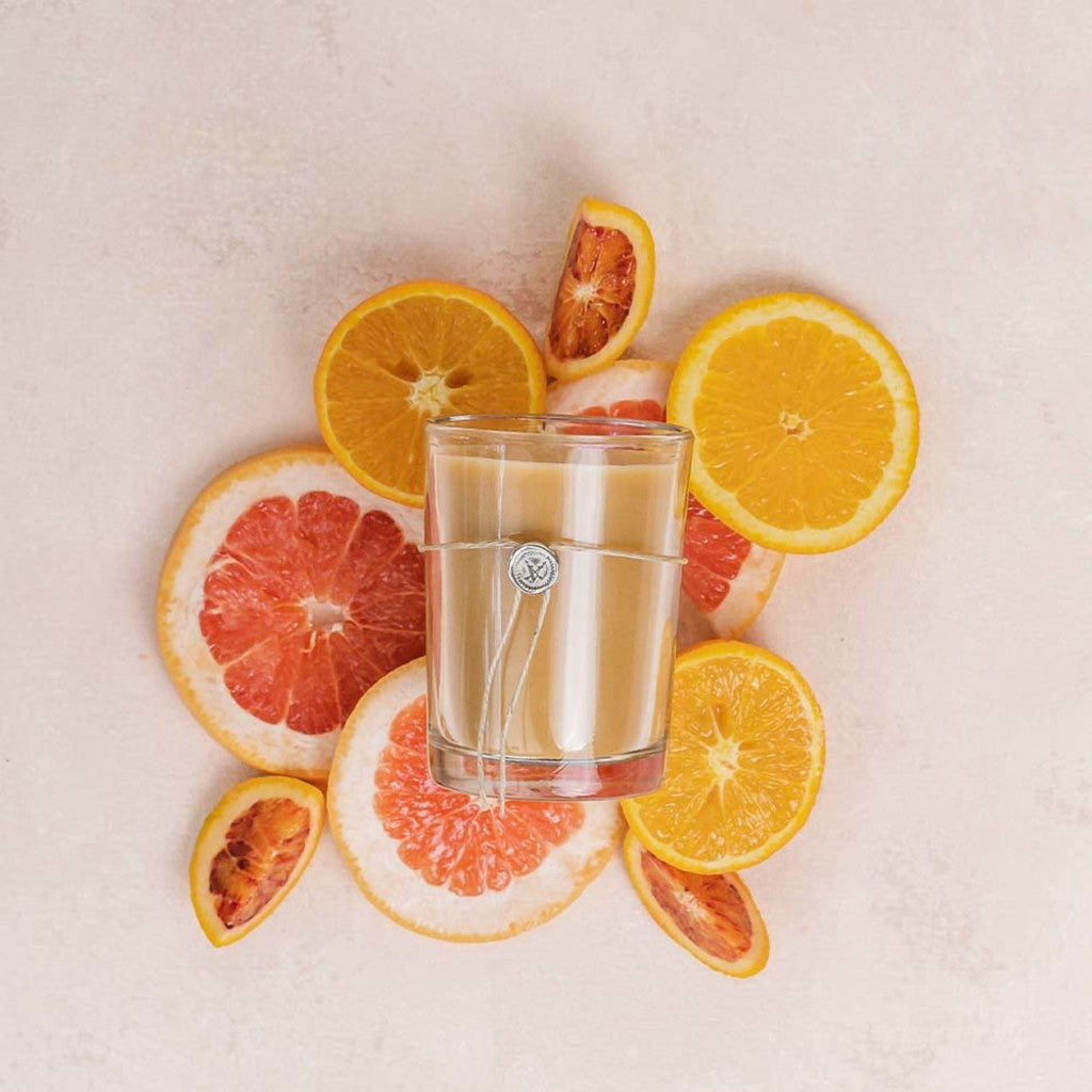 The Votivo Island Grapefruit 6.8 oz. Aromatic Candle, made with soy blend wax and featuring a silver charm, is displayed with slices of island grapefruit, orange, and blood orange on a light beige surface.