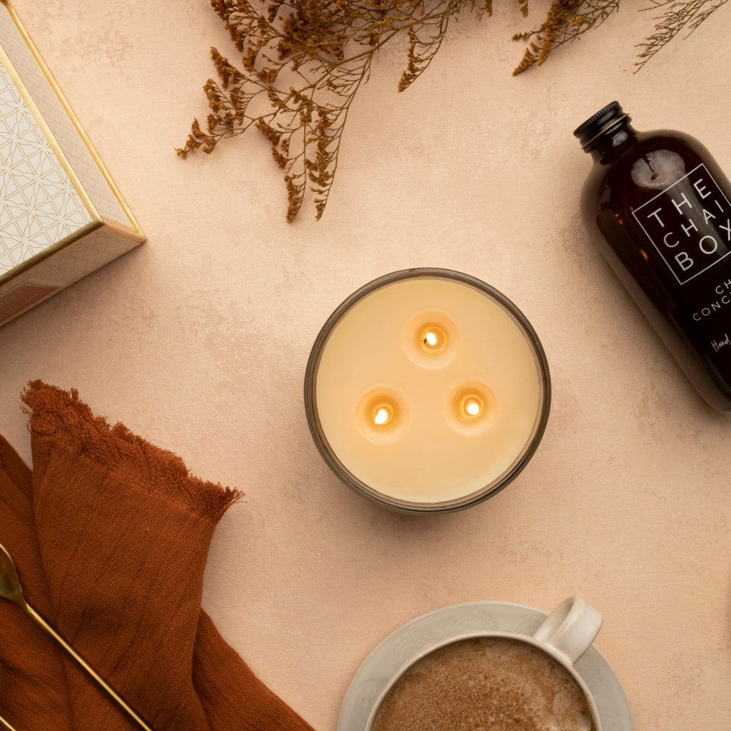 The Votivo Spiced Chai Holiday 3 Wick Candle, crafted from soy blend wax, glows on a beige surface beside coffee, dried flowers, a golden spoon, and THE CHAI BOX, filling the air with the cozy scent of spiced chai.