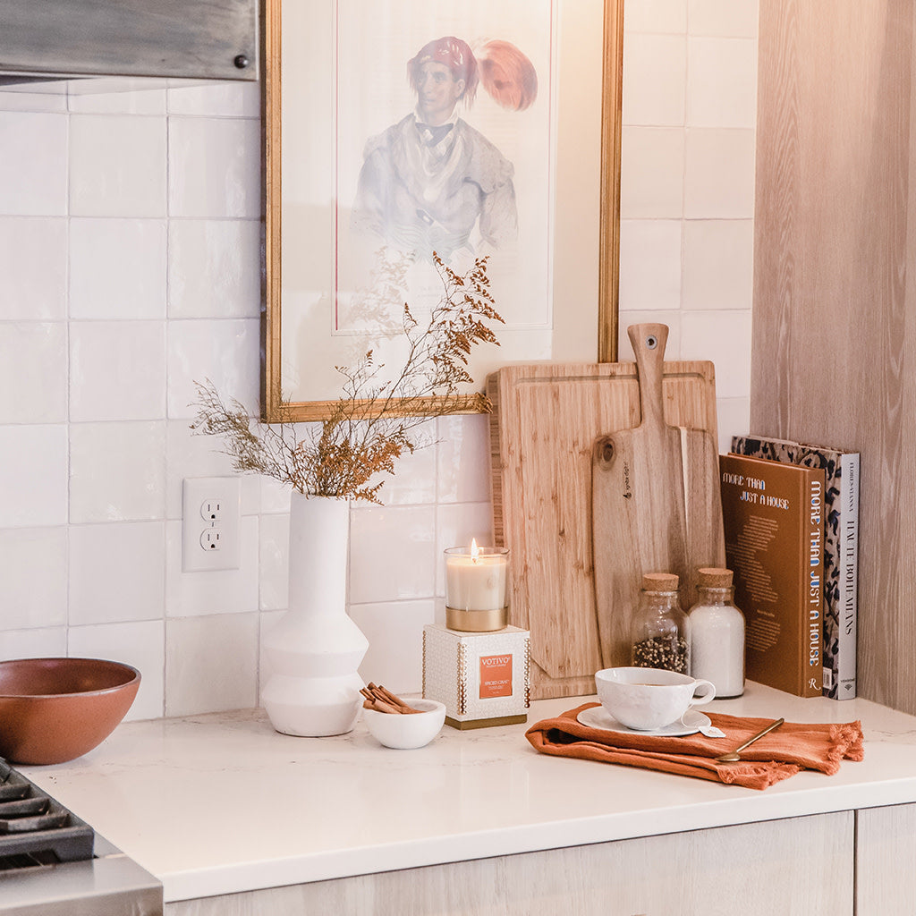 A cozy kitchen countertop displays cutting boards, a vase of dried flowers, a bowl, cup and saucer, salt and pepper shakers, stacked cookbooks, and the Votivo Spiced Chai 10 oz. Holiday Candle, set against tiled walls and framed artwork.