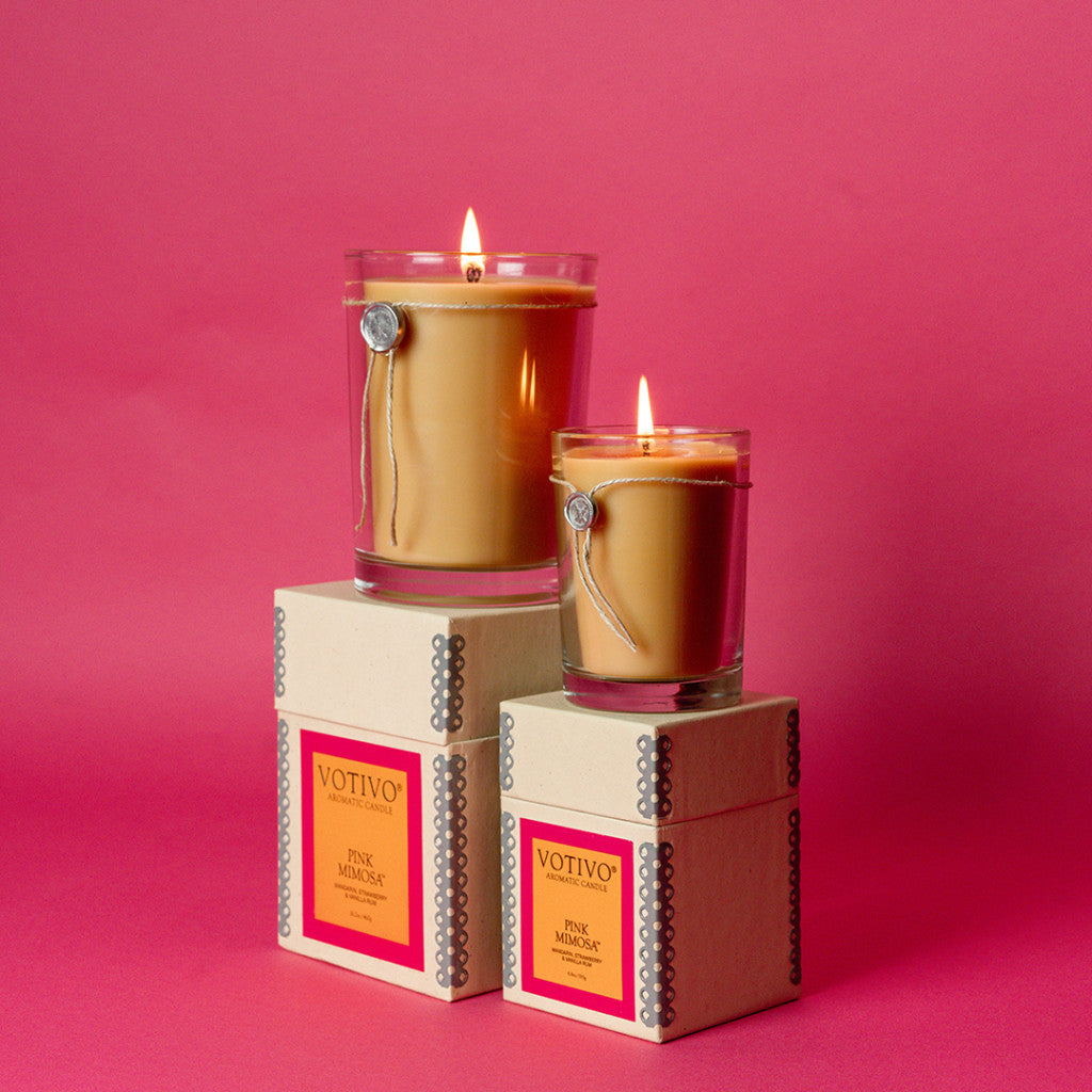 Two lit Votivo Pink Mimosa 6.8 oz. aromatic candles in clear jars rest on cream Pink Mimosa boxes against a pink background. Each soy blend candle features a metal charm tied around the rim and comes in different sizes.