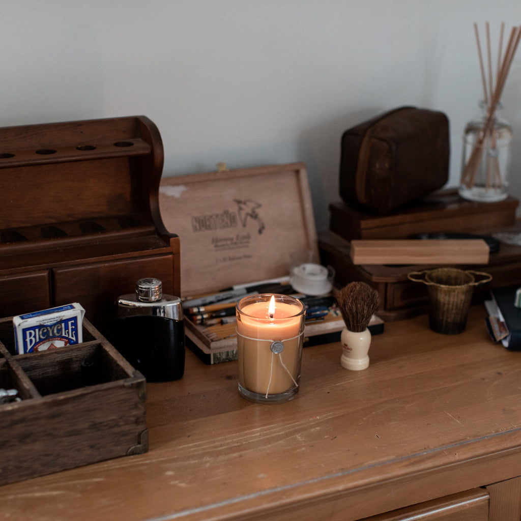 The Votivo Smoked Wood & Amber 6.8 oz. Aromatic Candle rests on a wooden desk, surrounded by a playing card deck, shaving brush, metal flask, wooden box, grooming tools, and small containers.