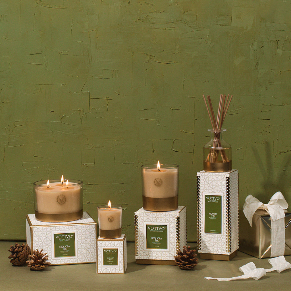 Five beige Votivo candles in glass jars and a Sequoia Fir Holiday Reed Diffuser by Votivo with rattan reeds are arranged on green and white boxes among pinecones and a gift box, against an olive green textured wall. All candles are lit.
